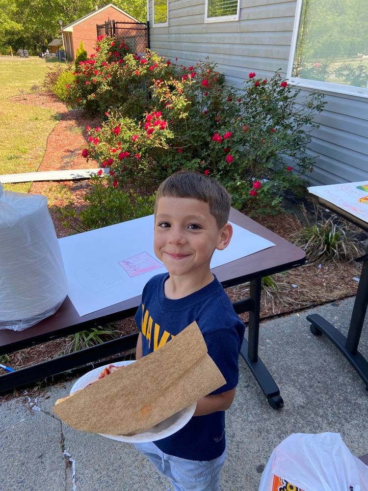 Little boy all smile outside holding a paper plate that has a slice of pizza